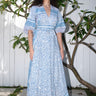 A woman standing on a pathway with a white and blue floral print maxi dress with three-quarter sleeves and a belt, wearing blue shoes.