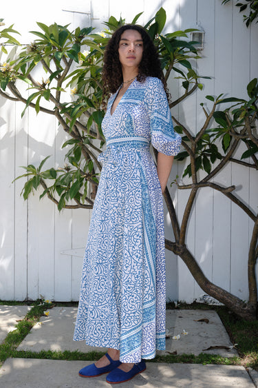 A woman standing on a pathway with a white and blue floral print maxi dress with three-quarter sleeves and a belt, wearing blue shoes.