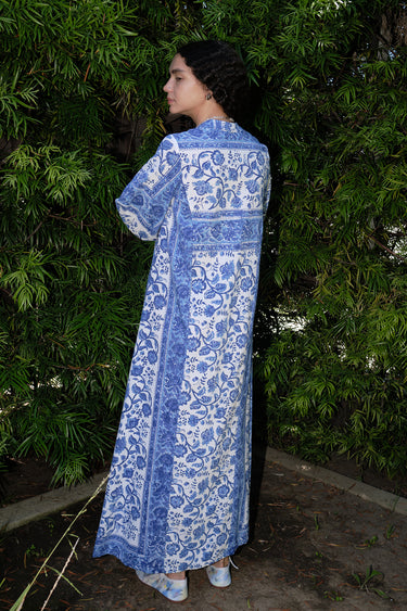A woman standing in a garden wearing a blue and white floral maxi dress with three quarter balloon sleeves and a V-neck back view.