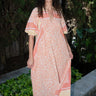 A woman standing in an outdoor setting wearing a maxi dress with orange and yellow floral patterns on a light background, featuring three quarter balloon sleeves and a relaxed, flowing cut.