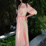 A woman standing in a garden wearing a pink and orange maxi dress with three-quarter sleeves and a belt, the dress is made of cotton hand block printed fabric.