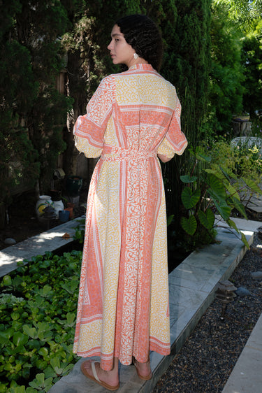 A woman standing in a garden wearing a pink and orange maxi dress with three-quarter sleeves and a belt, the dress is made of cotton hand block printed fabric.