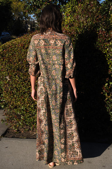 A woman wearing a maxi caftan with a floral pattern in a color that could be described as celadon green ground with tan, brown and blue, standing outdoors, back view.