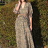 A woman wearing a maxi caftan with a floral pattern in a color that could be described as celadon green ground with tan, brown and blue, belted, standing outdoors.
