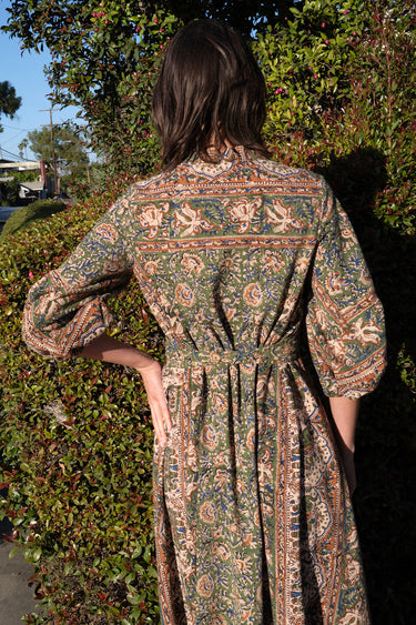 A woman wearing a maxi caftan with a floral pattern in a color that could be described as celadon green ground with tan, brown and blue, belted, standing outdoors, back view. 