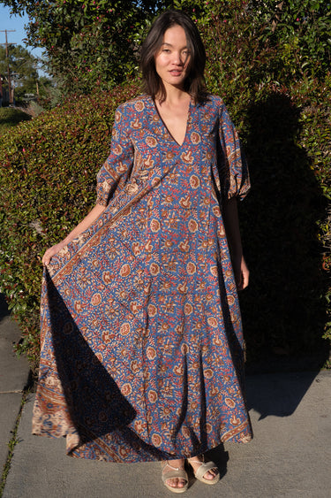 A woman wearing a maxi caftan with a floral pattern in a color that could be described as blue ground with tan, brown and coral, standing outdoors.