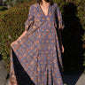 A woman wearing a maxi caftan with a floral pattern in a color that could be described as blue ground with tan, brown and coral, standing outdoors.