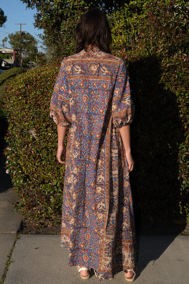 A woman wearing a maxi caftan with a floral pattern in a color that could be described as blue ground with tan, brown and coral, standing outdoors, back view. 