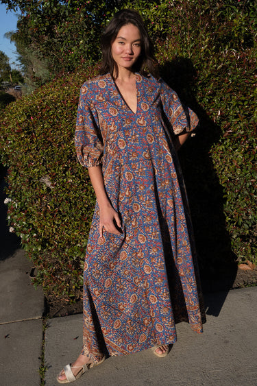 A woman wearing a maxi caftan with a floral pattern in a color that could be described as blue ground with tan, brown and coral, standing outdoors.