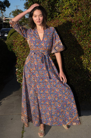 A woman wearing a maxi caftan with a floral pattern in a color that could be described as blue ground with tan, brown and coral, belted, standing outdoors.