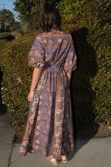A woman wearing a maxi caftan with a floral pattern in a color that could be described as blue ground with tan, brown and coral, belted, standing outdoors, back view.