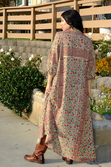 A woman standing outside wearing a beige ground with red and brown floral caftan. Back View