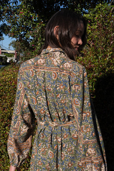 A woman wearing a green floral tapestry jacket with big patch pockets and an elastic shirred sleeve cuff, standing outside with greenery. 