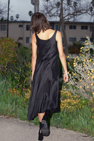 A woman standing on a pathway wearing a sleeveless, full-length black dress with a full drape neckline, side zipper, and pockets.  back view