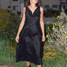A woman standing on a pathway wearing a sleeveless, full-length black dress with a full drape neckline, side zipper, and pockets. She has boots on and is facing away from the camera.