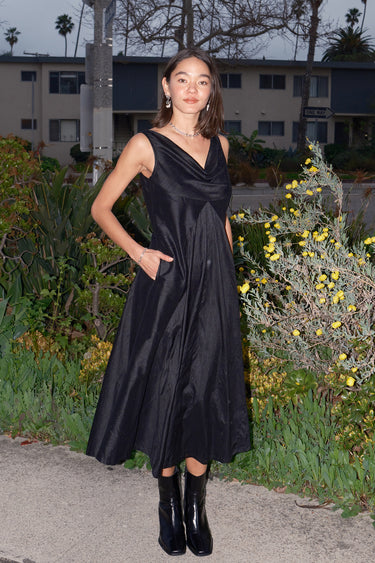 A woman standing on a pathway wearing a sleeveless, full-length black dress with a full drape neckline, side zipper, and pockets. She has boots on.