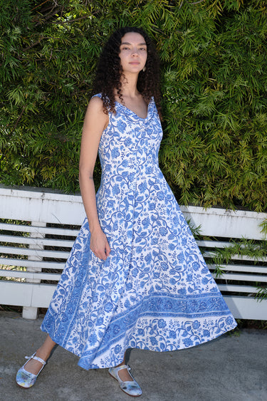 Woman standing in front of white fence and hedge wearing a white with blue tapestry midi dress with a draped neck and  very full skirt.