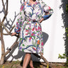 A woman standing outdoors wearing a multicolored coat dress with a floral tapestry pattern, featuring a tie belt and patch pockets.