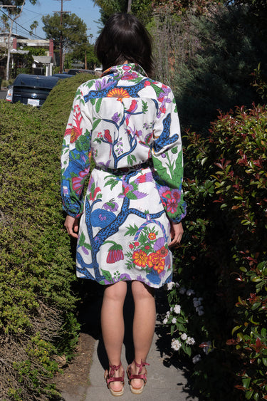 A woman standing on a sidewalk wearing a white coat dress with a colorful floral tapestry pattern, featuring patch pockets and a tie belt, back view.
