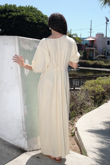 A woman standing outside by a canal wearing an alabaster off white maxi dress with shape from princess panels, back view. 