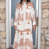 A woman standing in a doorway wearing a double-breasted Cypress Tapestry Jacket with Indian tapestry patterns in beige and brown colors with matching dress.