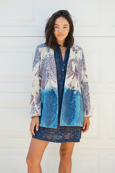 A woman smiling, wearing a double-breasted jacket with an indigo and blue pattern jacket, over a dress in a dark blue tapestry pattern. The jacket is dipped in indigo dye and has pockets on the inside.
