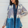 A woman smiling, wearing a double-breasted jacket with an indigo and blue pattern jacket, over a dress in a dark blue tapestry pattern. The jacket is dipped in indigo dye and has pockets on the inside.