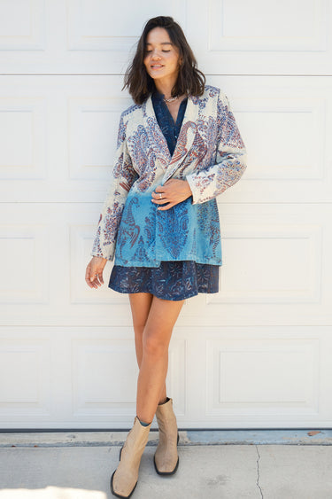 A woman smiling, wearing a double-breasted jacket with an indigo and blue pattern jacket, over a dress in a dark blue tapestry pattern. The jacket is dipped in indigo dye and has pockets on the inside.