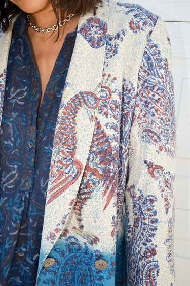 Detail of a woman wearing a double-breasted jacket with an indigo and blue pattern jacket, over a dress in a dark blue tapestry pattern. The jacket is dipped in indigo dye and has pockets on the inside.