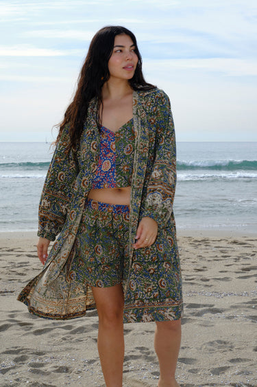 A woman standing at the beach wearing a floral tapestry low rise short cut using both green and blue tapestry of the same print tapestry mixed together features front pockets. Worn with a matching camisole and coat dress.