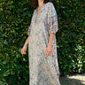 A female model wearing a multicolored floral caftan with three-quarter sleeves and standing in front of a hedge.