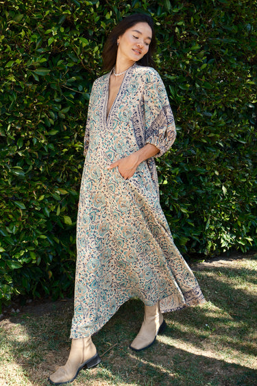 A female model wearing a multicolored floral caftan with three-quarter sleeves and standing in front of a hedge.