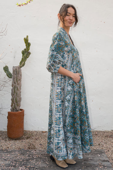 Woman in a patterned Caftan standing outdoors with a white wall and cactus in the background.