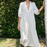 A woman standing outdoors wearing a white caftan with 3/4 balloon sleeves and a deep 'V' neckline. The caftan has pockets on the front panel and is made of a non-sheer crinkle textured fabric.