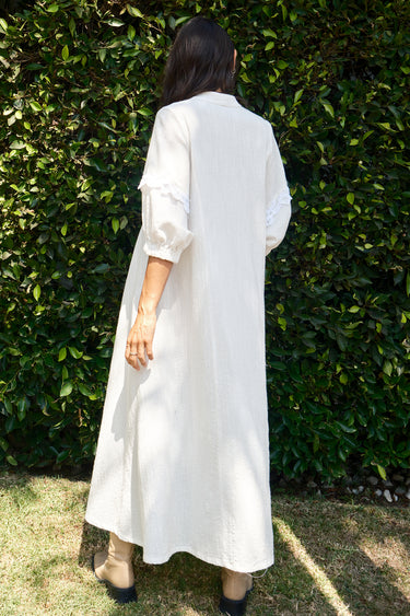 A woman standing outdoors wearing a white caftan with 3/4 balloon sleeves and a deep 'V' neckline. The caftan has pockets on the front panel and is made of a non-sheer crinkle textured fabric. Back View