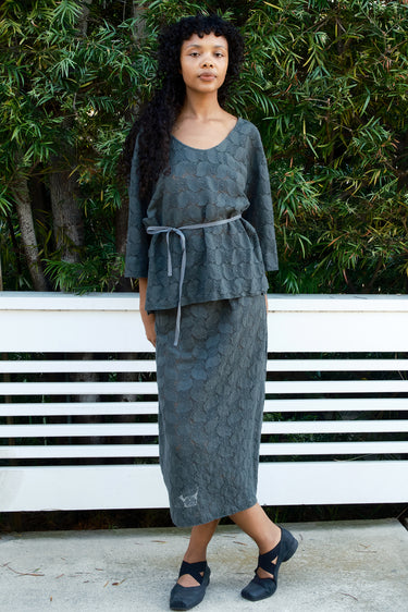 Woman wearing a gray textured outfit standing outdoors with greenery in the background