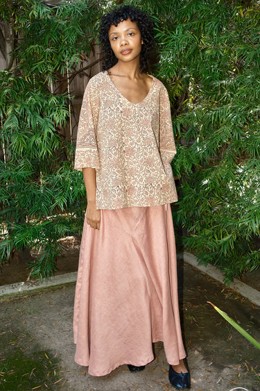 Woman wearing a pink lace top and skirt standing in front of green foliage.