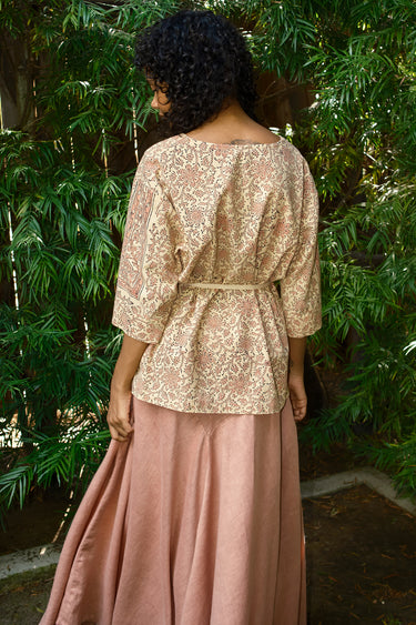 Woman wearing a patterned top and pink skirt standing in front of green foliage