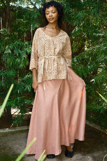 Woman wearing a beige top and pink skirt standing in front of green foliage.