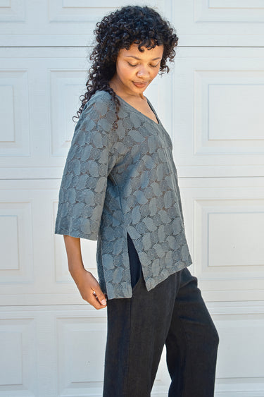 Woman wearing a patterned gray top and black pants against a white wall.