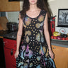 A woman standing in a kitchen wearing a knee-length dress in a black ground tapestry with rainbow  suns, moons and stars printed on it. 
