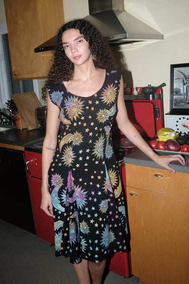 A woman standing in a kitchen wearing a knee-length dress in a black ground tapestry with rainbow  suns, moons and stars printed on it. 
