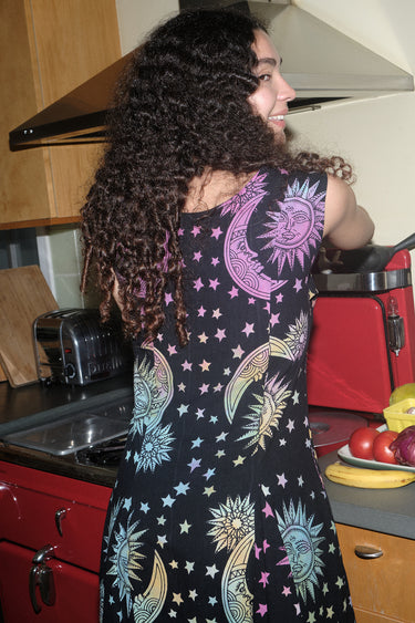 A woman standing in a kitchen wearing a knee-length dress in a black ground tapestry with rainbow  suns, moons and stars printed on it, back view. 