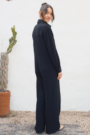 Person wearing a black jumpsuit standing against a white wall with a cactus plant.