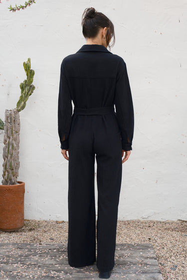 Person wearing a black jumpsuit standing against a white wall with a cactus plant.