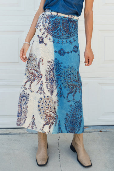 A woman standing in front of a white garage wearing a blue and beige peacock printed tapestry skirt with a denim knot top. Tan ankle boots 