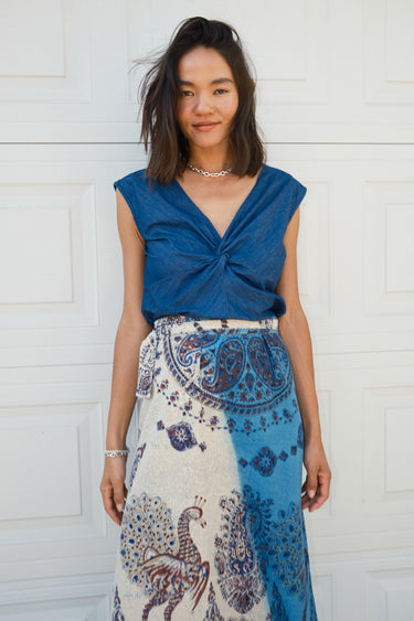 A woman standing in front of a white garage wearing a blue and beige peacock printed tapestry skirt with a denim knot top. 