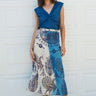 A woman standing in front of a white garage wearing a blue and beige peacock printed tapestry skirt with a denim knot top. Tan ankle boots 