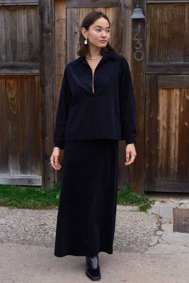 A woman standing in front of a brown wooden fence , wearing a long, black, corduroy skirt with a V-neck top.