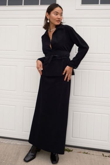 A woman standing in front of a white garage door, wearing a long, black, corduroy skirt with a V-neck top.